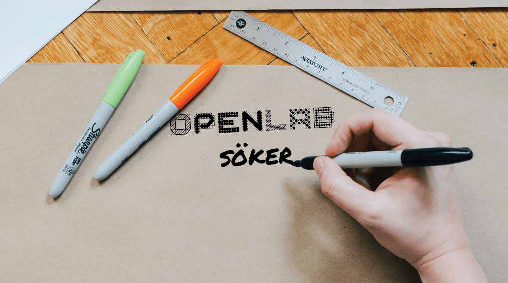 En hand skriver "Openlab söker" med en svart penna på gråbrunt papper.