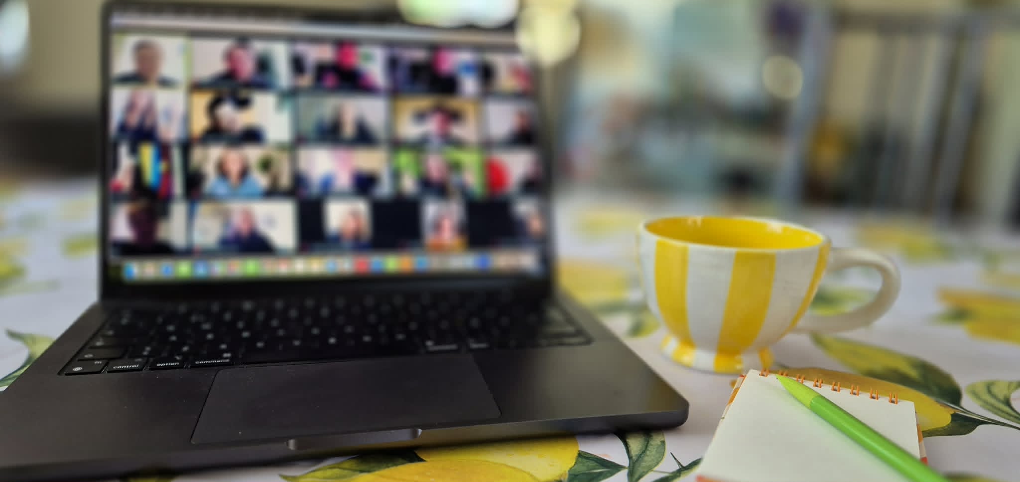 En bärbar dator visar ett pågående videomöte med flera deltagare, bredvid en gul- och vitrandig kaffekopp på en citronmönstrad duk.