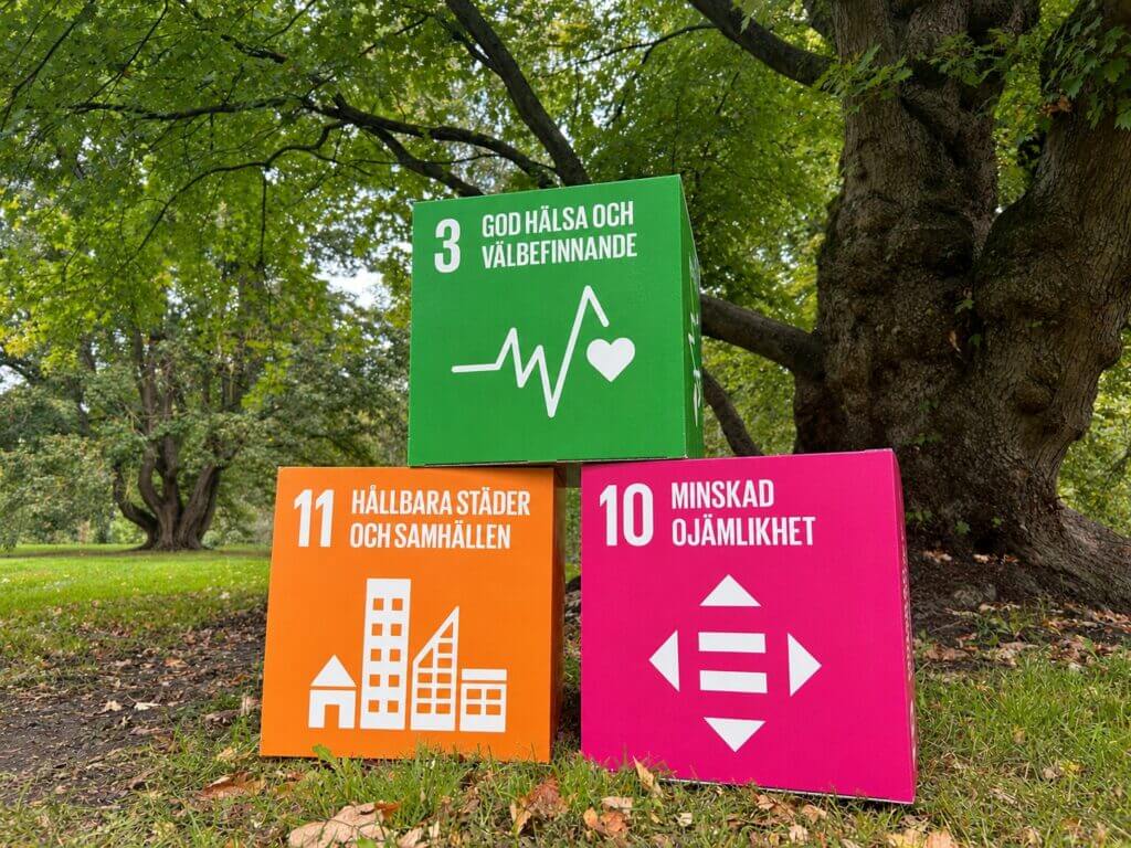 Tre färgglada kuber som symboliserar de globala hållbarhetsmålen nr 3, 10 och 11, placerade i en lummig park.